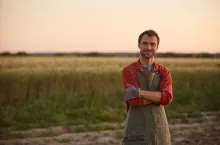 Waist up portrait of young farmer posing confidently with arms crossed while standing in field at sunset and smiling at camera, copy space