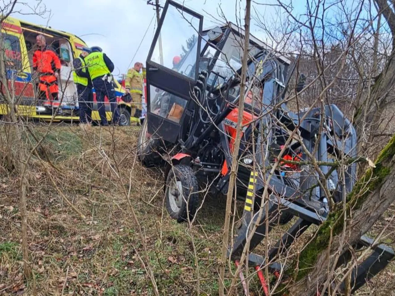 Czerwony ursus poza drogą oparty przodem o drzewo, a na drodze powyżej znajduje się karetka pogotowia